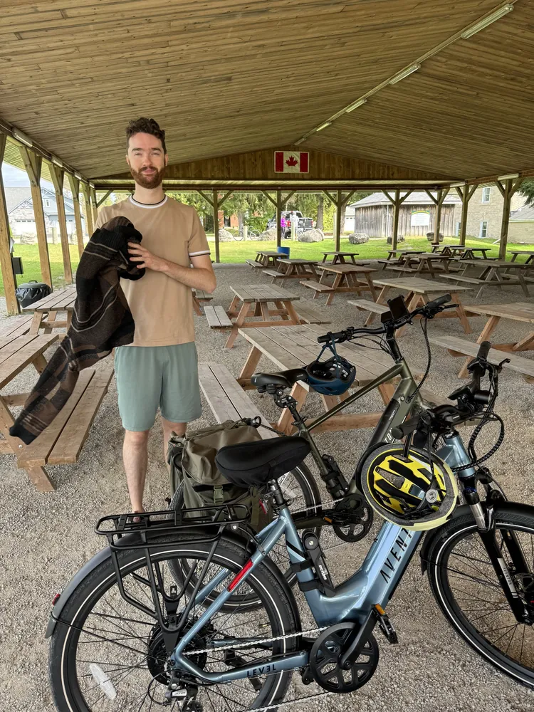 Skyler standing under a picnic pavilion beside two electric bikes with helmets on the handlebars.