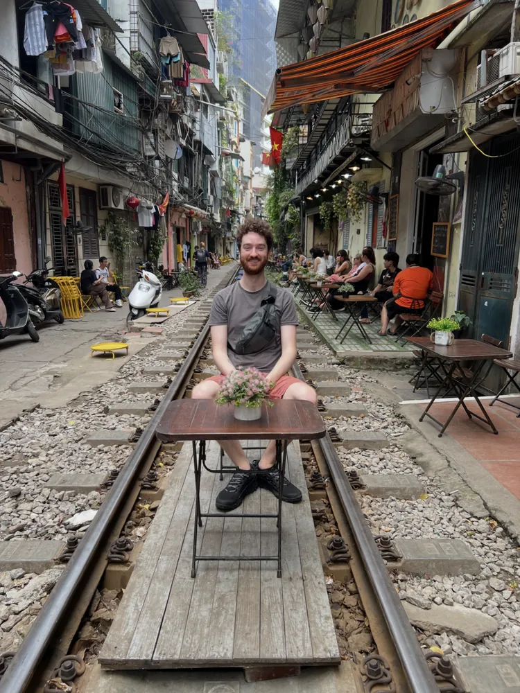 Skyler sitting at a small café table set directly on active train tracks in a narrow Hanoi street.