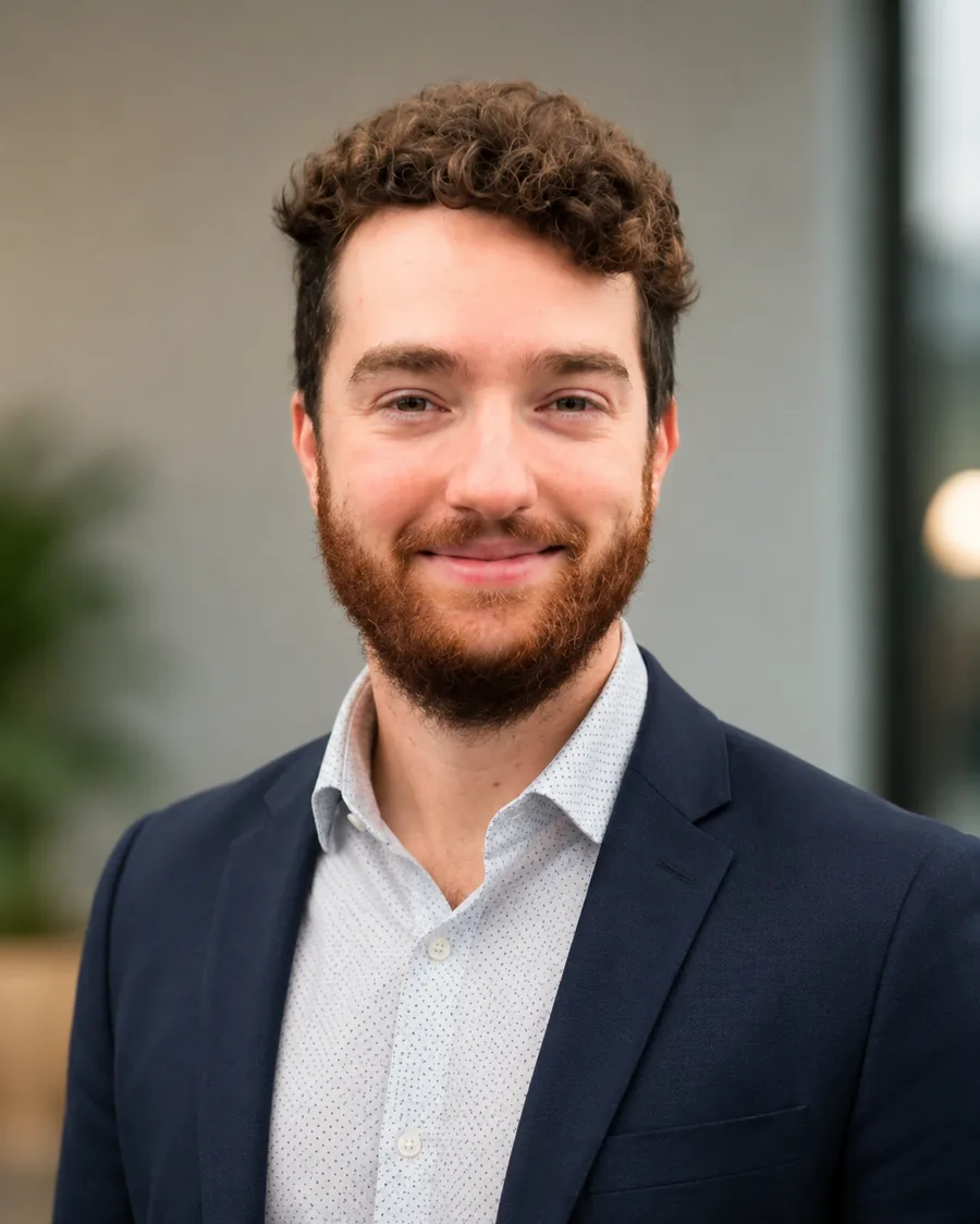 Portrait of Skyler Hammond – smiling, curly brown hair and a red-brown beard, wearing a navy blazer over a white dotted shirt.