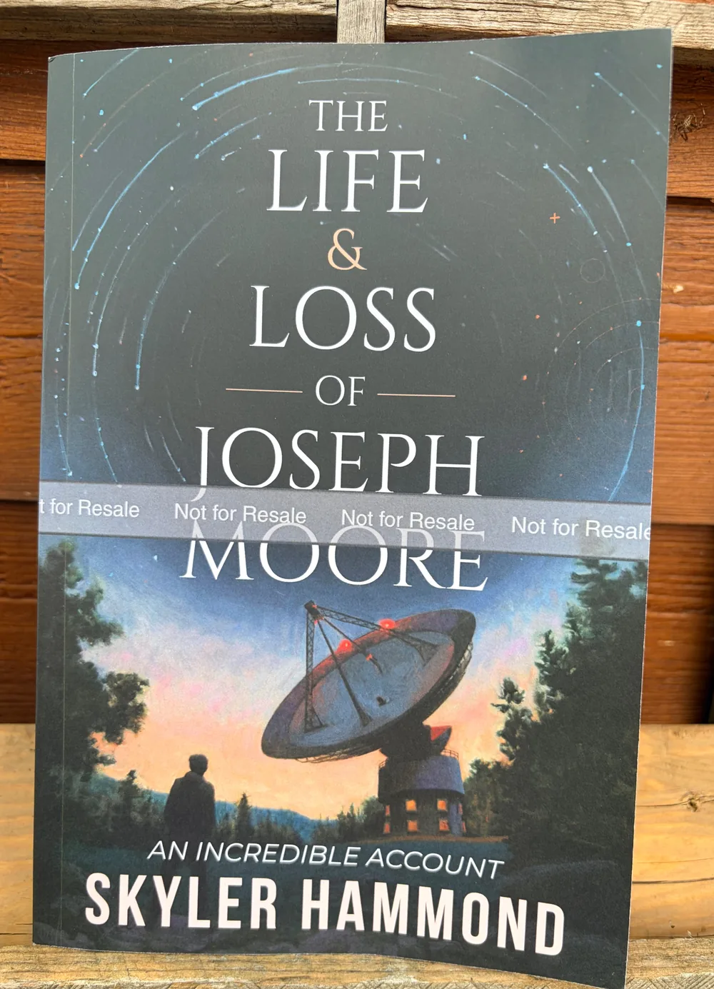 Paperback copy of The Life & Loss of Joseph Moore by Skyler Hammond, propped against a wooden wall. The cover shows a figure at dusk looking toward a radio telescope under a sky of star trails.