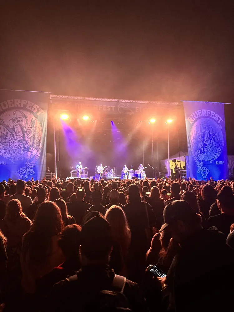 A festival crowd at night facing a stage lit in orange and purple, flanked by Riverfest Elora banners.