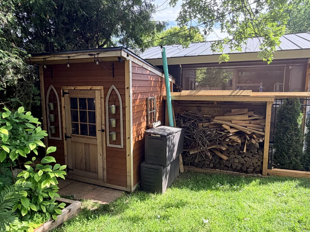A backyard garden shed with dark siding and a lean-to wood shed stacked with firewood beside it.